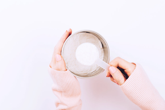 A hand in a pink sweater holding a scoop of white collagen powder over a container, symbolizing a measured supplement intake.